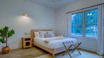 A bedroom with a bed, bedside table, lamp, potted plant, and a window with a view of the outside.