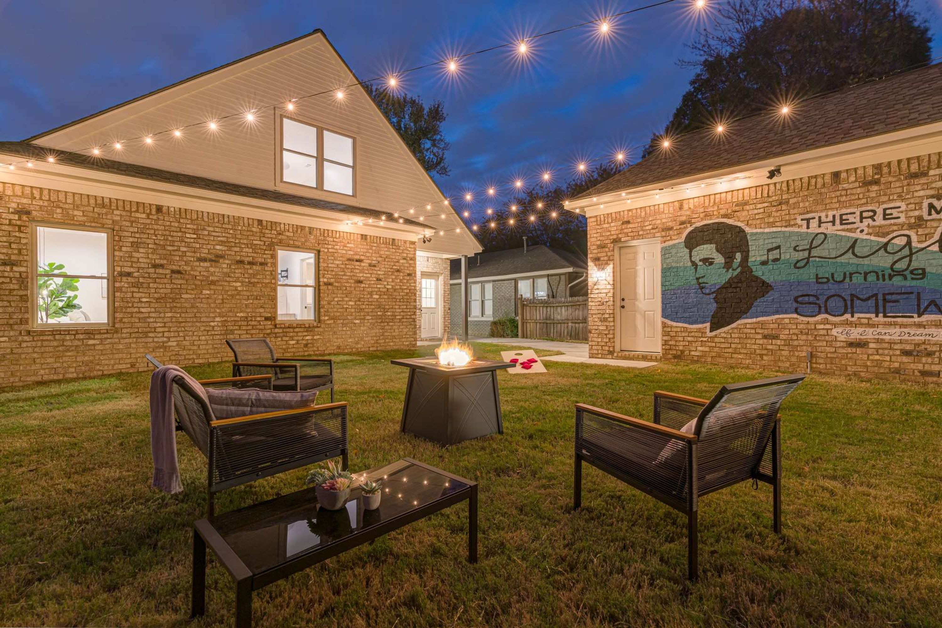 A backyard with a brick house, outdoor seating, a fire pit, and string lights.