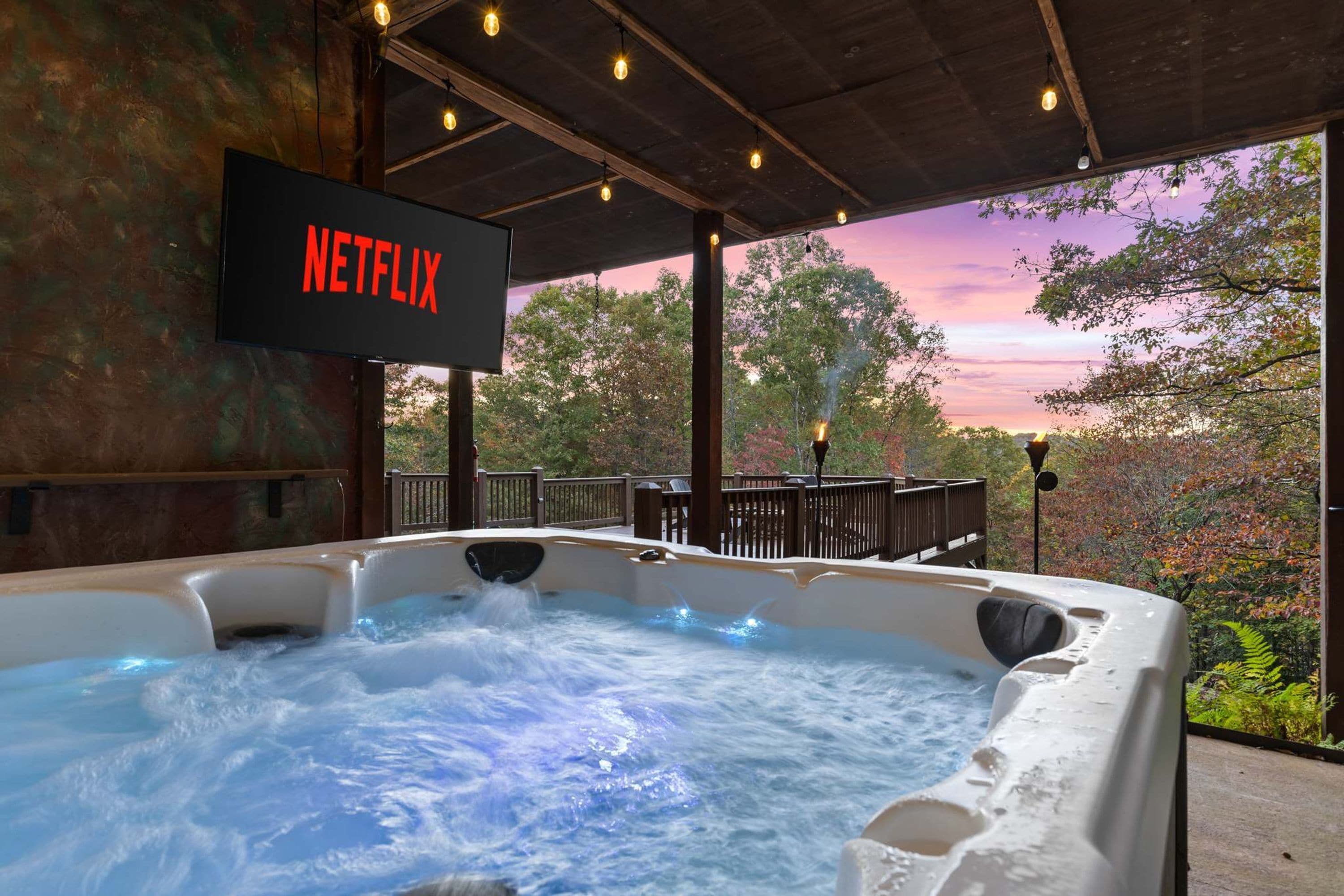 Un jacuzzi con vista a árboles y un atardecer, un cartel que dice 'NETFLIX' y una terraza de madera.