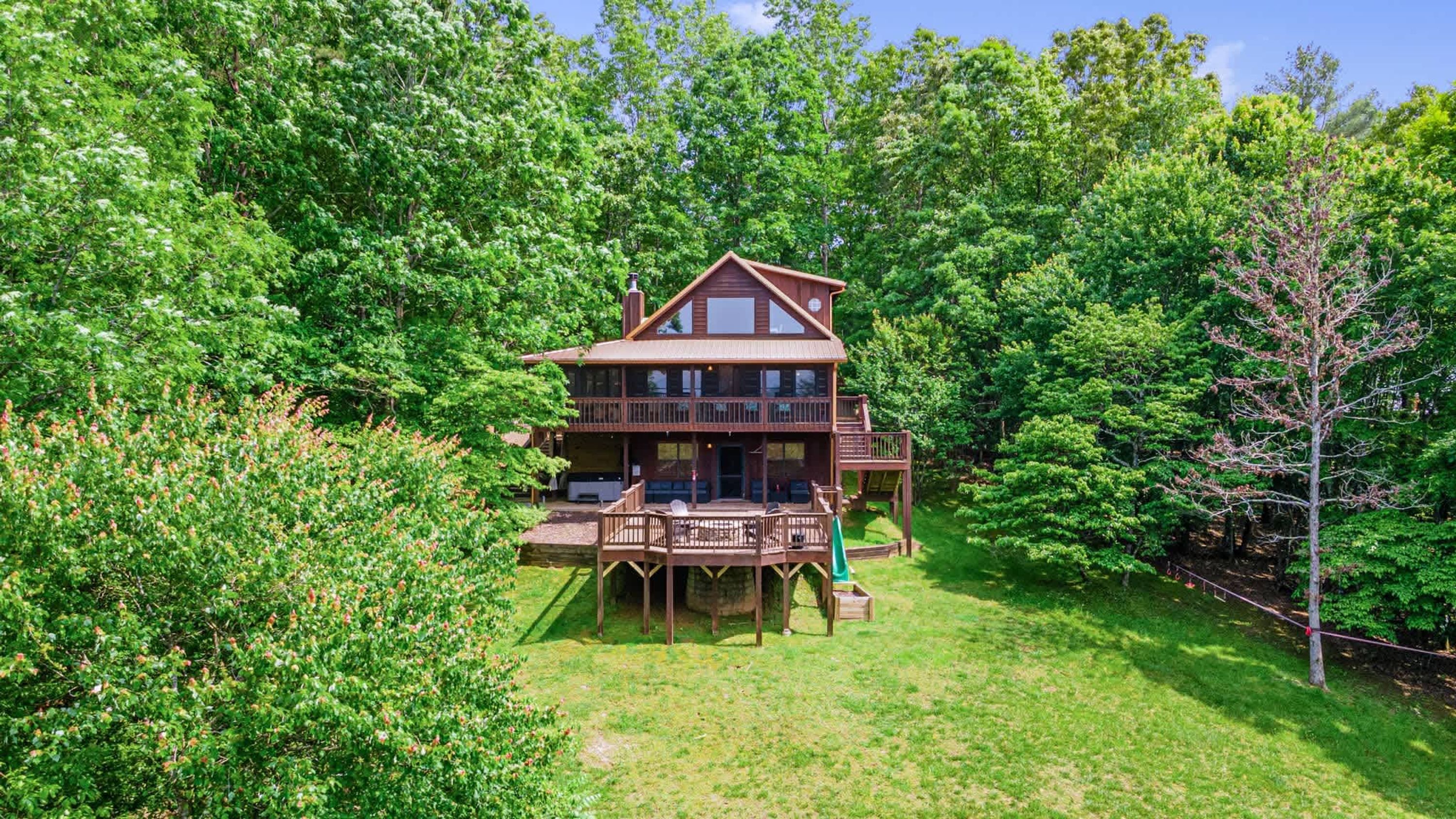 A two-story wooden cabin with a deck and a covered outdoor seating area, surrounded by dense green forest.