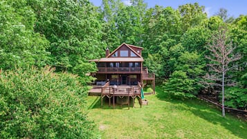 A two-story wooden cabin with a deck and a covered outdoor seating area, surrounded by dense green forest.