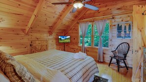A wooden cabin bedroom with a bed, a television, a chair, and a window with curtains. - Mountain Views w Slide Hot Tub Game Room (Mineral Bluff)