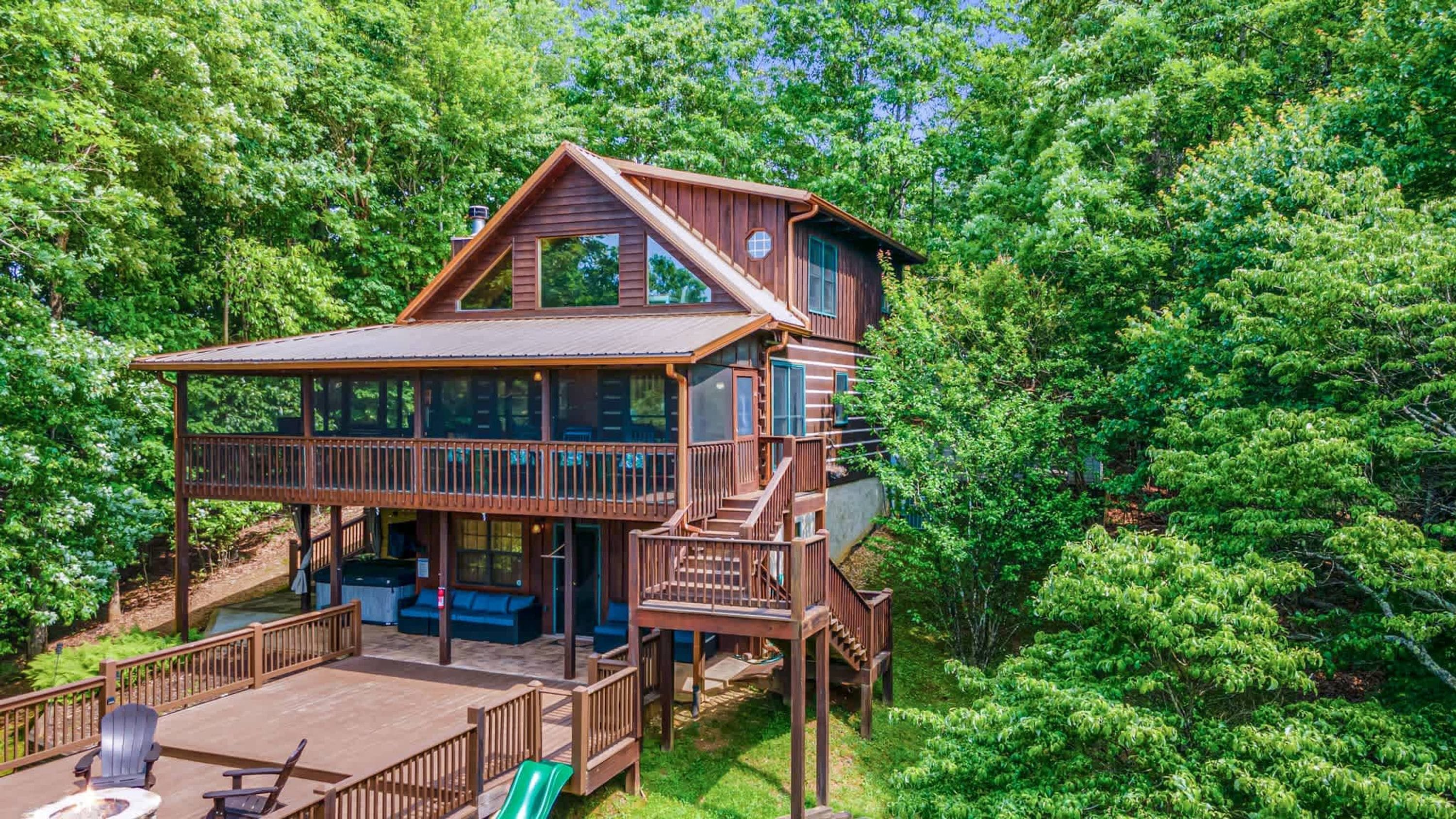 A multi-level wooden cabin with a deck, surrounded by dense forest.