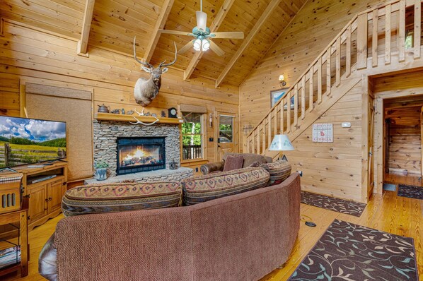 A cozy cabin interior with a stone fireplace, a ceiling fan, and a mounted deer head. - Mountain Views w Slide Hot Tub Game Room (Mineral Bluff)