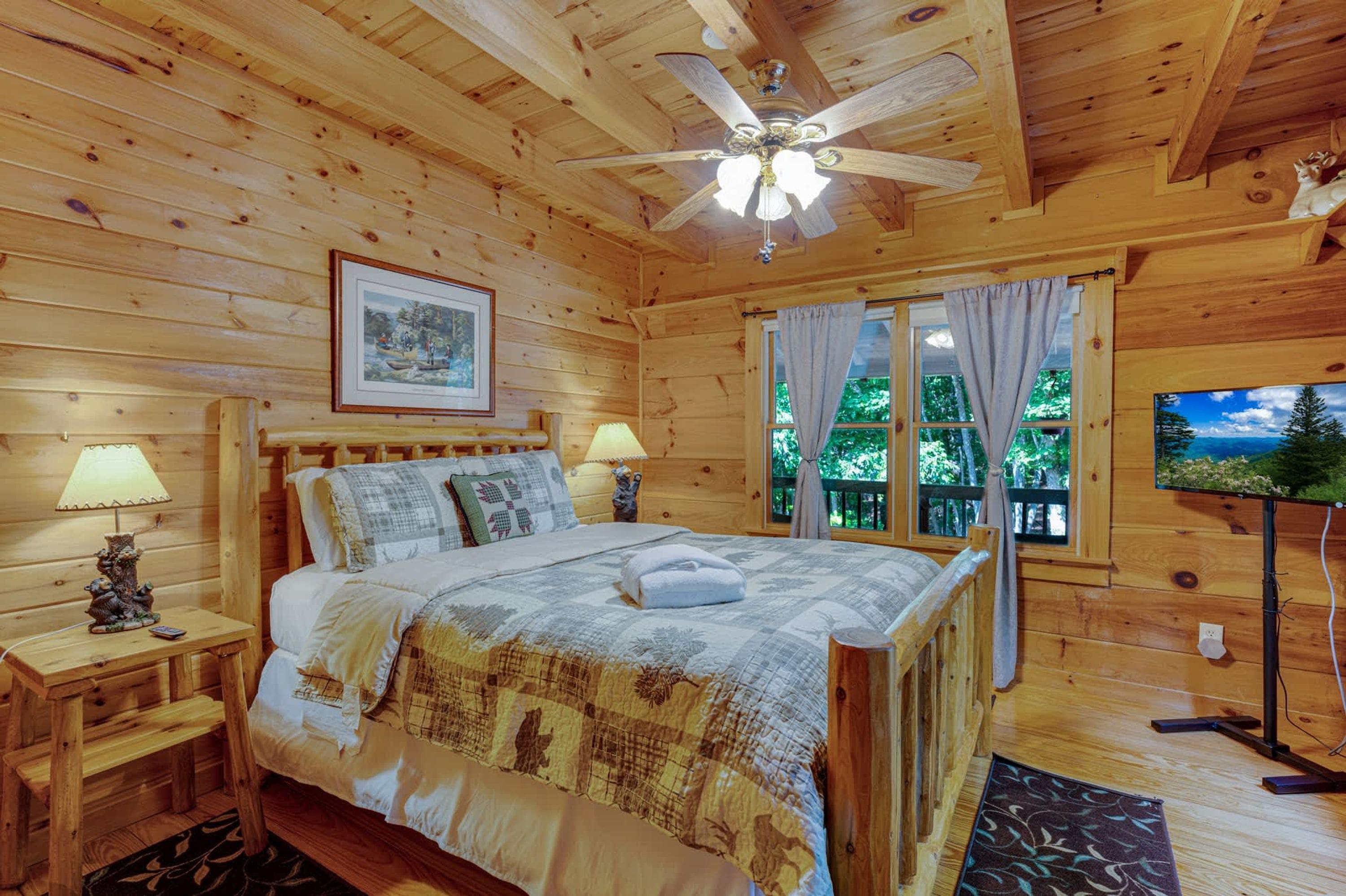Quarto de cabana de madeira com uma cama, duas luminárias de cabeceira, um ventilador de teto, um quadro emoldurado, uma janela com cortinas e uma televisão.