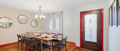 Un comedor con una mesa de madera dispuesta para cuatro personas, sillas alrededor, un papel tapiz decorado y una puerta con marco rojo.