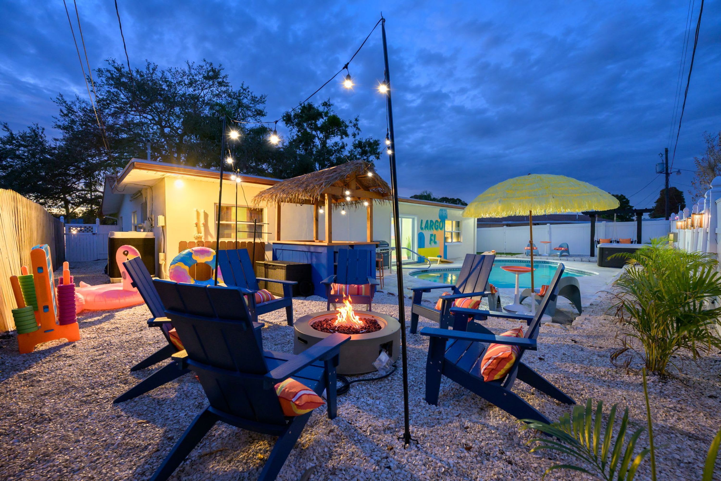 Un espace extérieur aménagé avec des chaises et un foyer, entouré d’une piscine et d’une structure à toit de chaume.