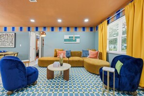 Un salon avec un sol à carreaux, un ensemble de canapés, deux fauteuils bleus, une petite table basse et une fenêtre avec des rideaux jaunes.