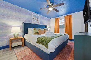 Una habitación de hotel con cama, mesita de noche, ventilador de techo, televisor y ventana con cortinas.