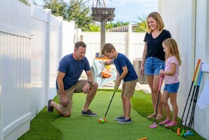 Une famille joue au croquet dans un jardin avec une pelouse artificielle et une clôture blanche.