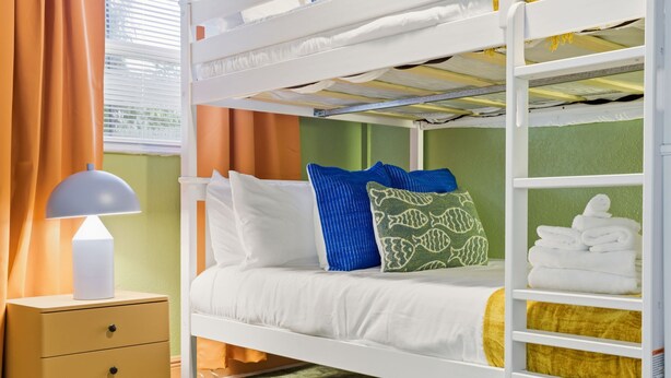 A bunk bed with white bedding, a bedside table with a lamp, and a window with curtains. - Backyard Oasis Pool Hot Tub Putt Putt Bar (Largo)