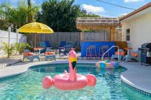 Una piscina con juguetes inflables, sillas de descanso y una sombrilla de paja.
