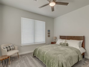 A bedroom with a bed, a chair, a nightstand, a ceiling fan, and a window with blinds. - House at the Refuge - Relax Take in Nature (Medicine Park)