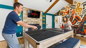 Una mesa de ping pong en una habitación con pared de piedra, un televisor mostrando 'NETFLIX' y un mural abstracto colorido.