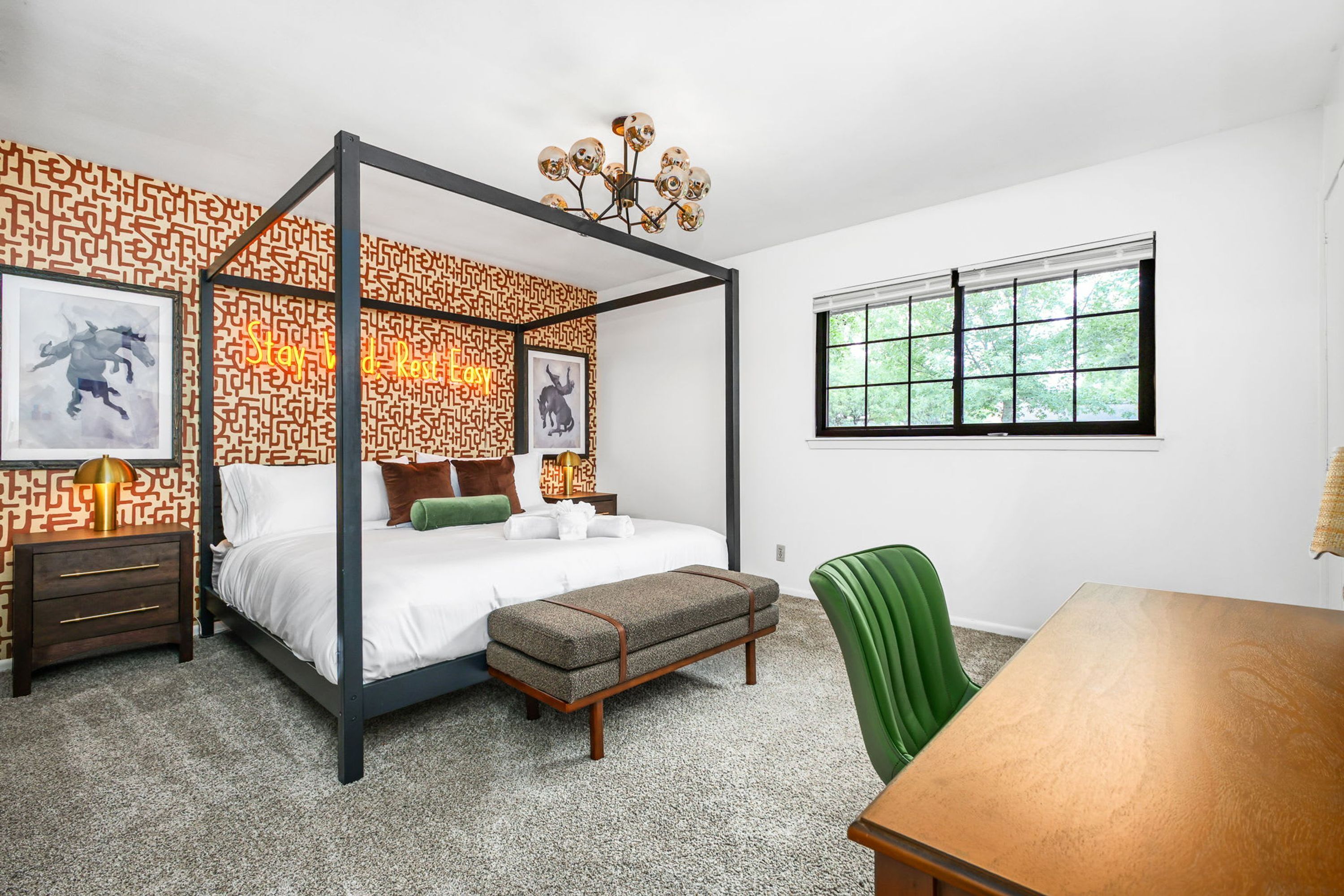 A four-poster bed with a geometric patterned wall, a green upholstered bench, a wooden table, and a window with a view of trees.