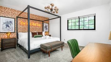 A four-poster bed with a geometric patterned wall, a green upholstered bench, a wooden table, and a window with a view of trees.