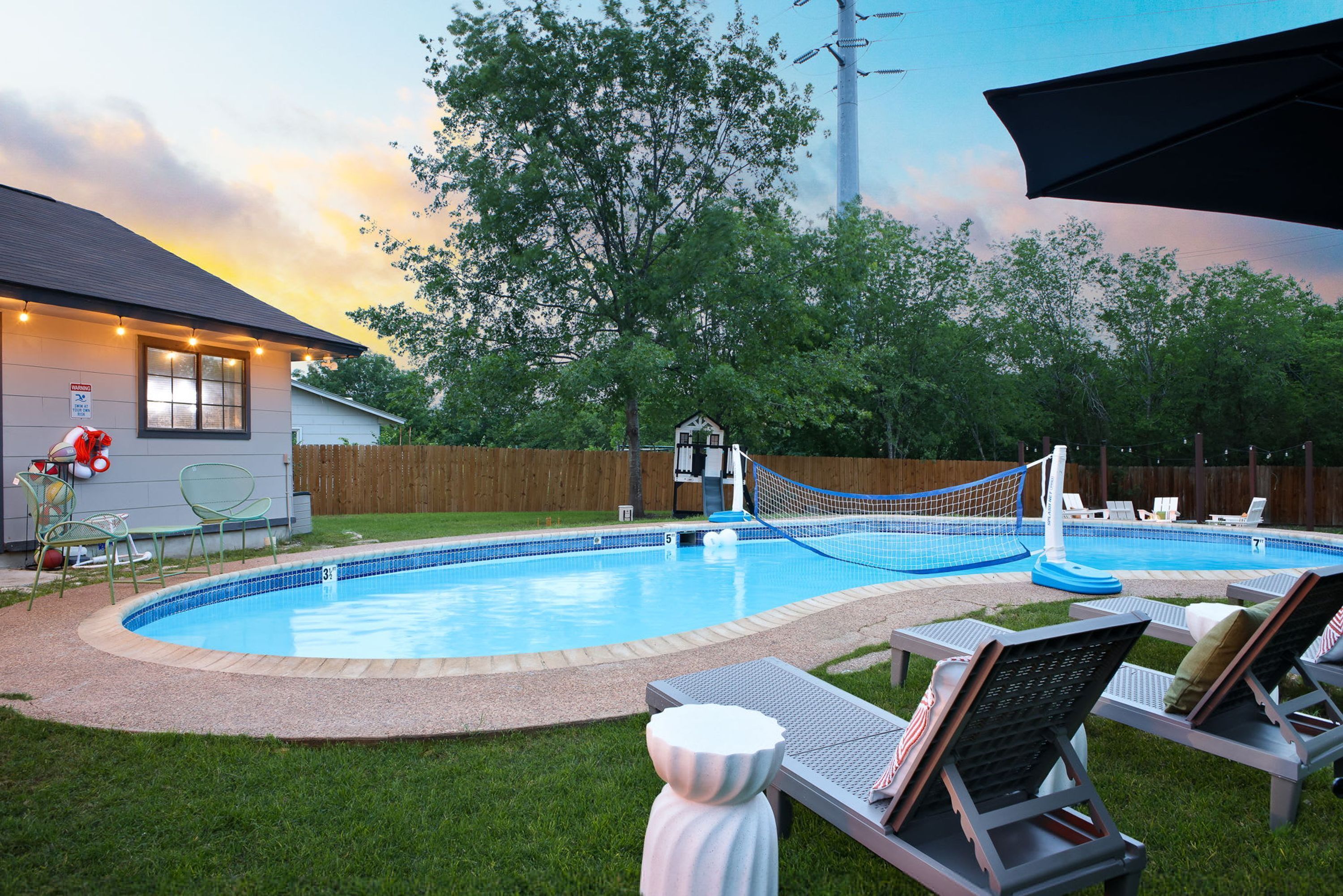 A backyard with a swimming pool, lounge chairs, a wooden deck, and a house with string lights.