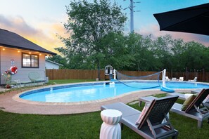 Un patio trasero con piscina, sillas de descanso, una terraza de madera y una casa con luces colgantes.