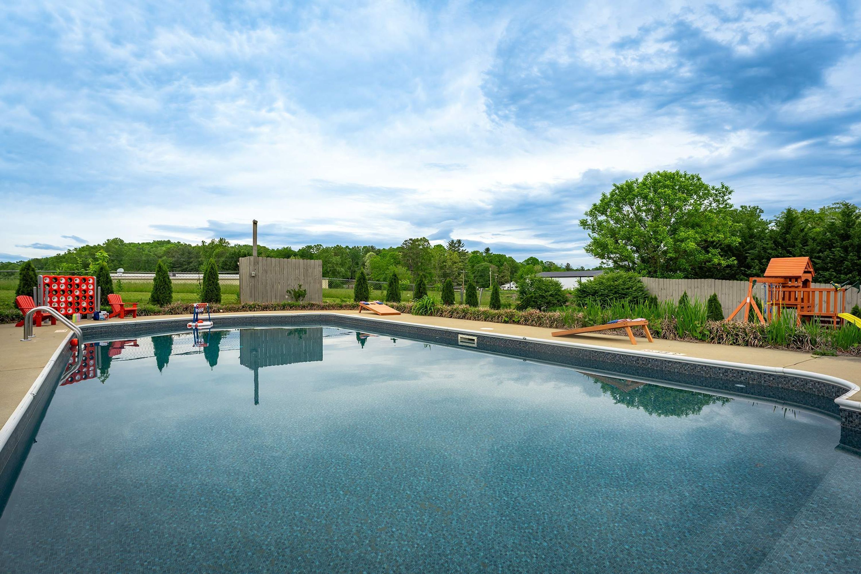 A clear swimming pool with a wooden deck and lounge chairs, surrounded by a fenced area with trees and a wooden play structure.
