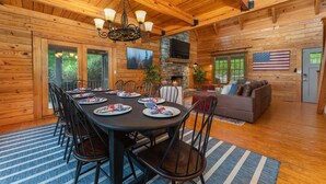 A wooden cabin interior with a dining area, a stone fireplace, and a living room with a couch and an American flag. - 7 Br Sleeps 20 Sport Court Pool Hot Tub Playground (Arden)