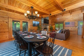 A wooden cabin interior with a dining area, a stone fireplace, and a living room with a couch and an American flag. - 7 Br Sleeps 20 Sport Court Pool Hot Tub Playground (Arden)