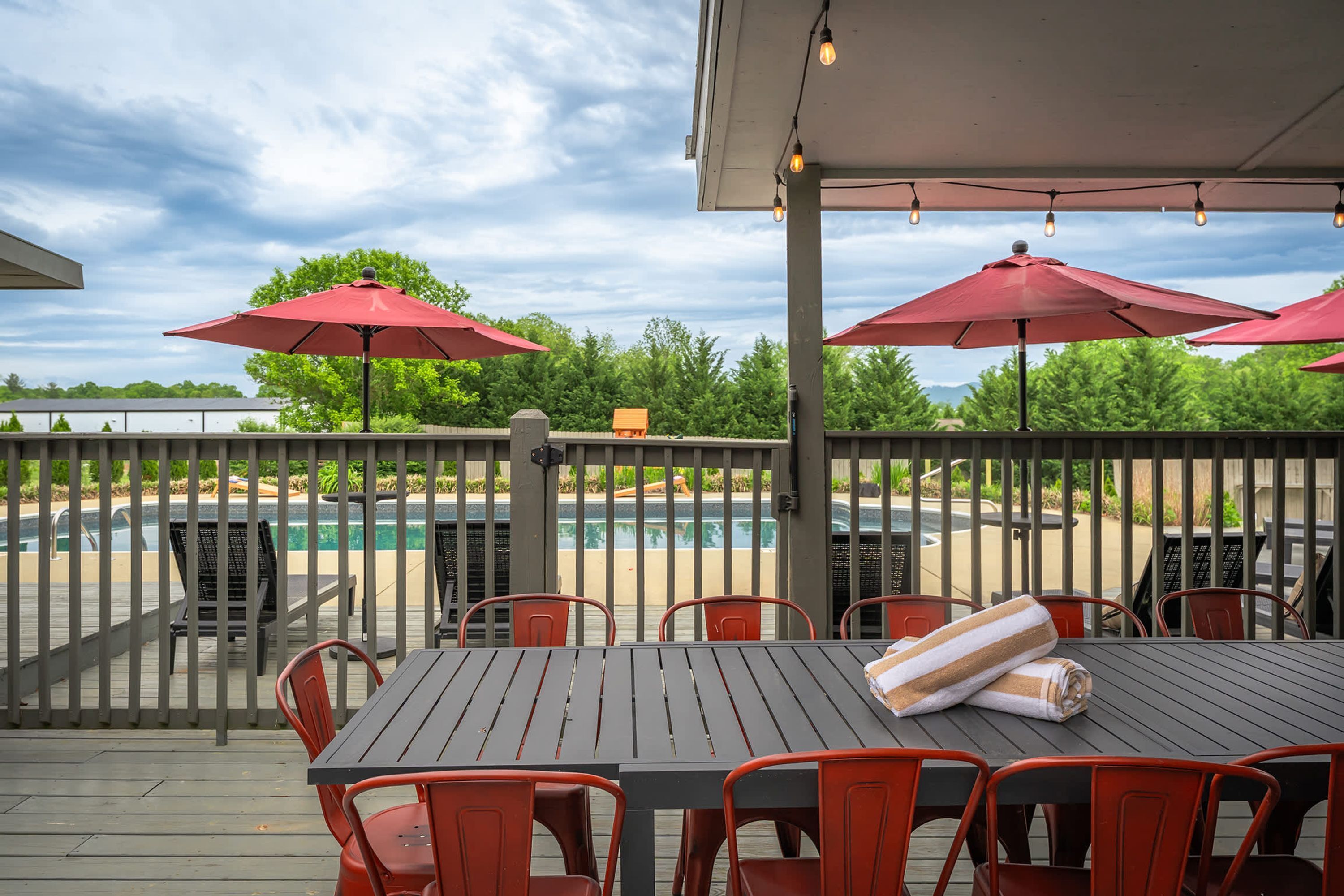 Une terrasse extérieure avec vue sur la piscine, agrémentée de parasols rouges, de chaises en métal et d’une table avec une serviette.