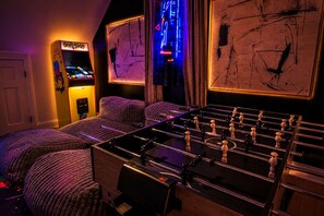 A room with a foosball table, two beds, a Pac-Man arcade machine, and wall art.