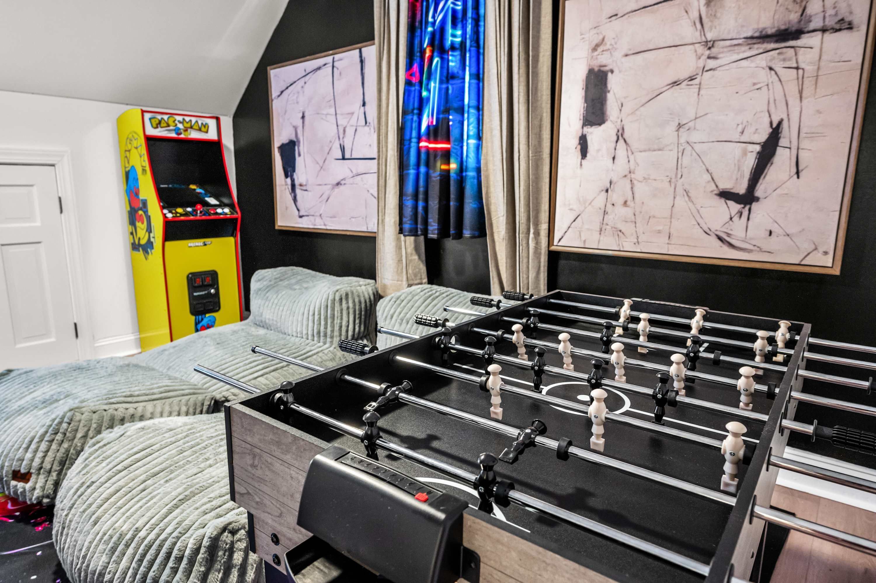 A room with a foosball table, a Pac-Man arcade machine, and abstract art on the walls.