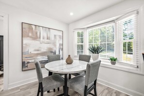 A dining area with a round table, four grey chairs, a vase, and a large abstract painting on the wall.