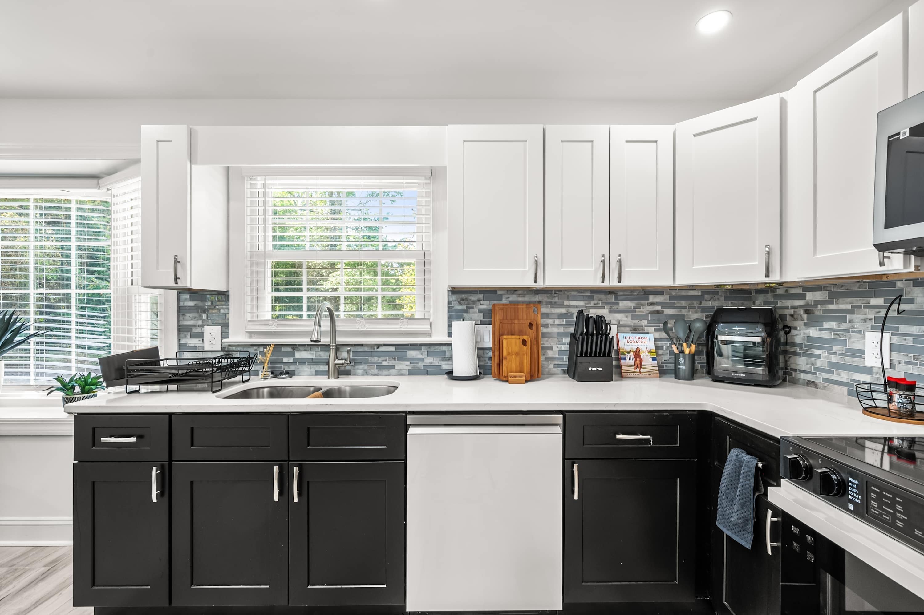 A modern kitchen with white and black cabinetry, a stainless steel sink, a built-in oven, and a large window with blinds.