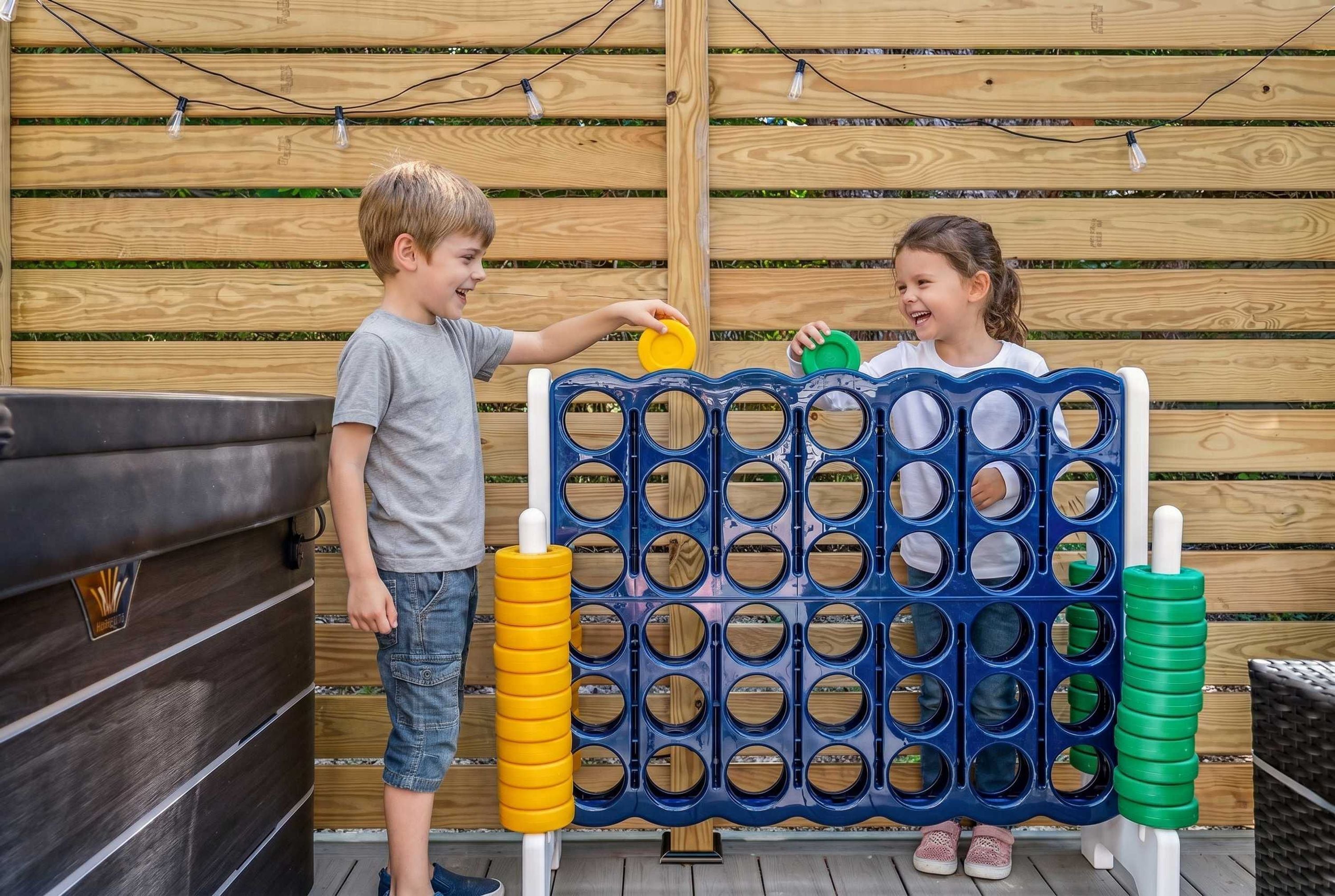 Dos niños jugando con un juego de mesa de madera, una parrilla y una guirnalda de luces en un patio trasero.