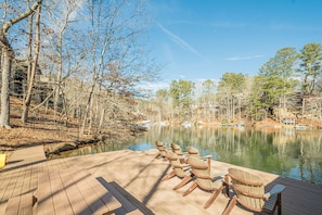 Une terrasse en bois, agrémentée de chaises Adirondack, surplombe un lac paisible, entouré d’arbres et de bâtiments.