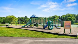 Ein Spielplatz mit mehreren Rutschen und Klettergerüsten, umzäunt und von Grünflächen umgeben.