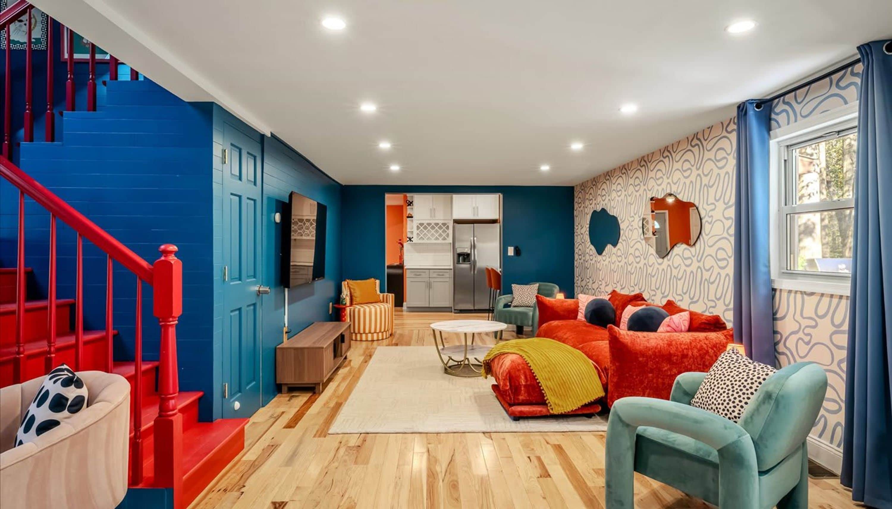 A modern living room with a blue staircase, a kitchen in the background, and a large window with curtains.