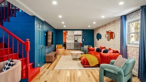 Un salon moderne avec un escalier bleu, une cuisine à l’arrière-plan et une grande fenêtre avec des rideaux.