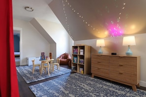 A cozy room with a sloped ceiling, a wooden cabinet, two lamps, a chair, and a small table. - Pine Paddle Getaway Game Room Hot Tub Pool (Muskegon)