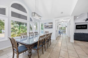 A spacious dining area with a large table and chairs, a television mounted on the wall, and a view of the outdoors through large windows. - Coastal Luxe Retreat - Heated Pool - Games - Games (Hilton Head Island)