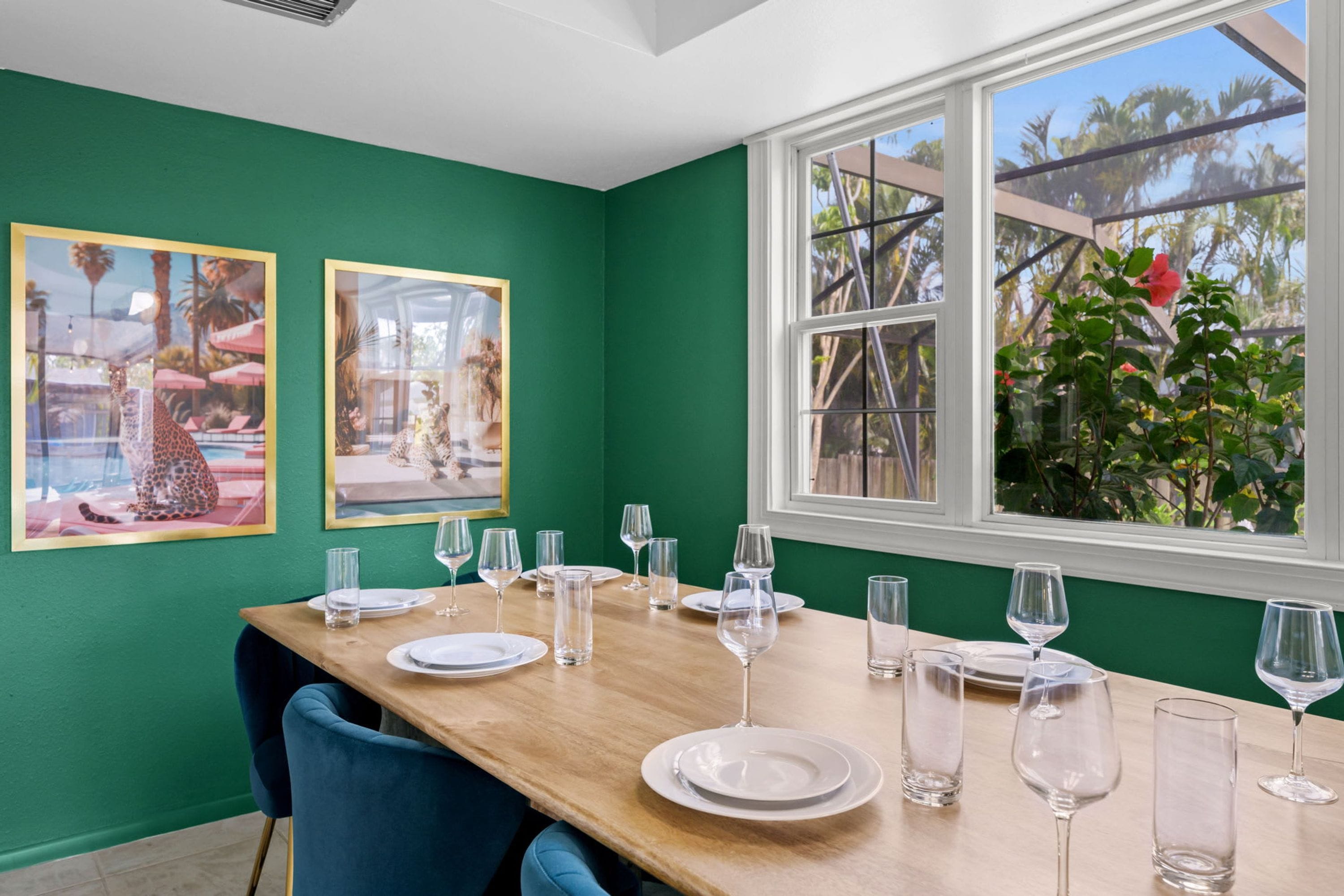 Un coin repas agrémenté d’un mur vert, de deux tableaux encadrés, d’une table en bois mise avec des assiettes et des verres, et d’une fenêtre donnant sur des palmiers.
