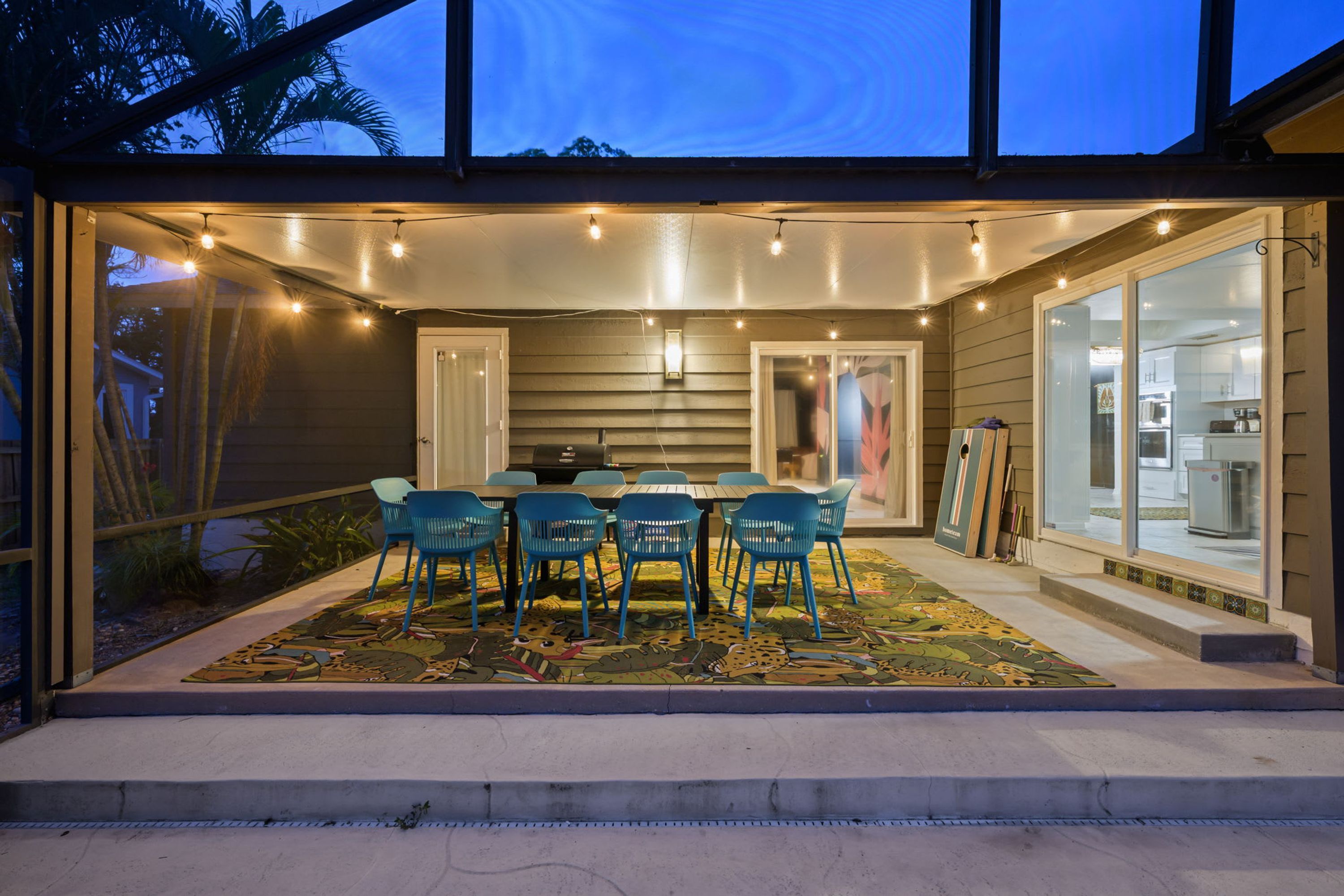 An outdoor dining area with a table and chairs, a patterned rug, and a view of the kitchen through glass doors.