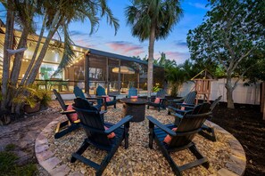 An outdoor seating area with Adirondack chairs and a fire pit, surrounded by a stone border and palm trees. - Family Retreat Pool Putt Putt Playground (Bradenton)