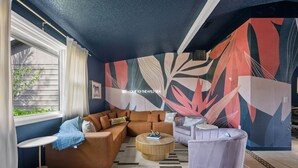 Ein modernes Wohnzimmer mit einer Eckcouch, einem runden Couchtisch und einem violetten Sessel. Eine große abstrakte Wandmalerei schmückt die Wand, und ein Fenster bietet Blick auf Grünflächen.