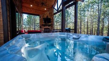 A hot tub in a cabin with a stone fireplace and a view of a forest.