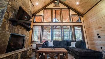 A log cabin interior with a stone fireplace, a wooden ceiling, and a large window.