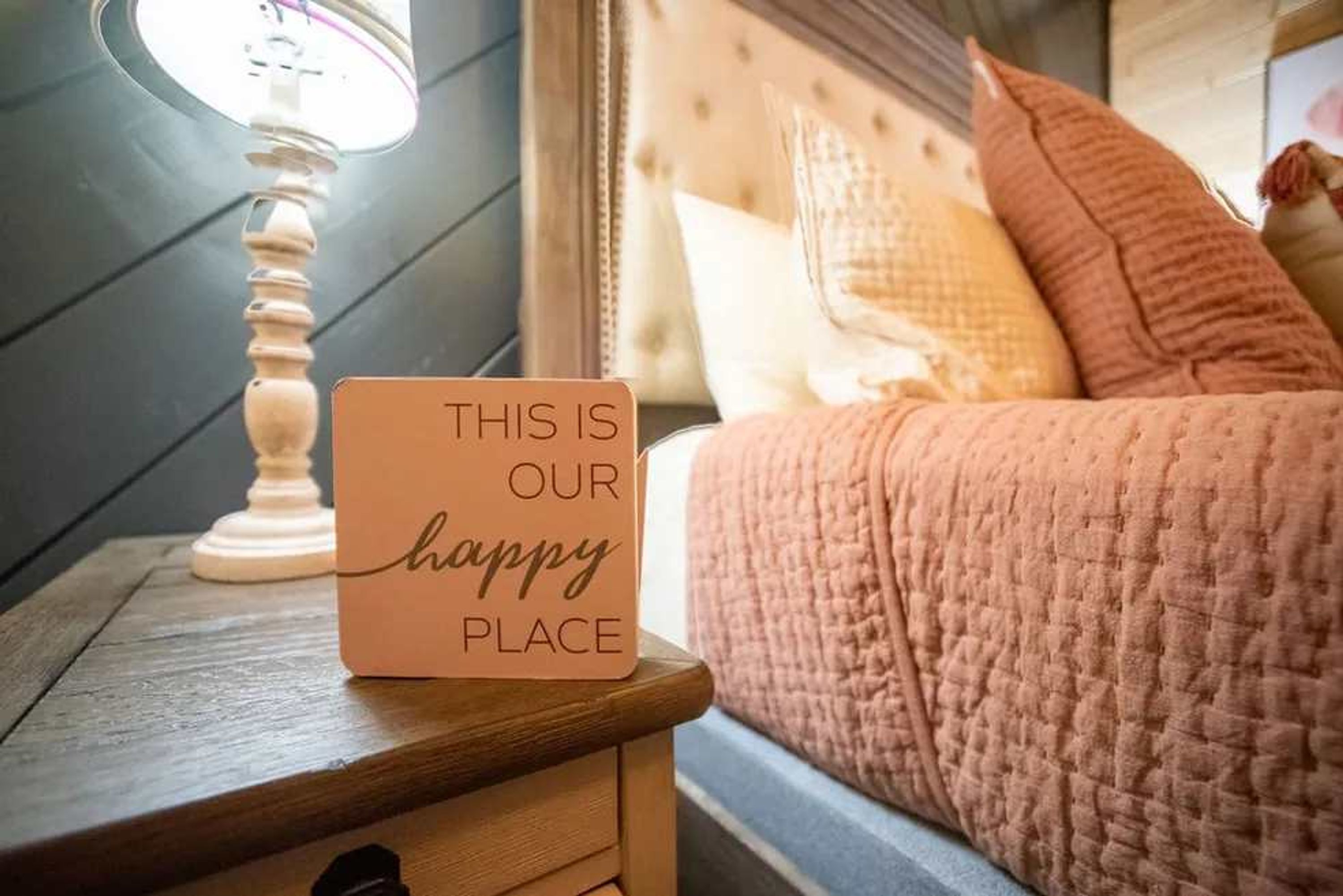 Ett sängbord med en lampa, en säng med ett rosa överkast och en skylt med texten: ”DET HÄR ÄR VÅR LYCKLIGA PLATS”.