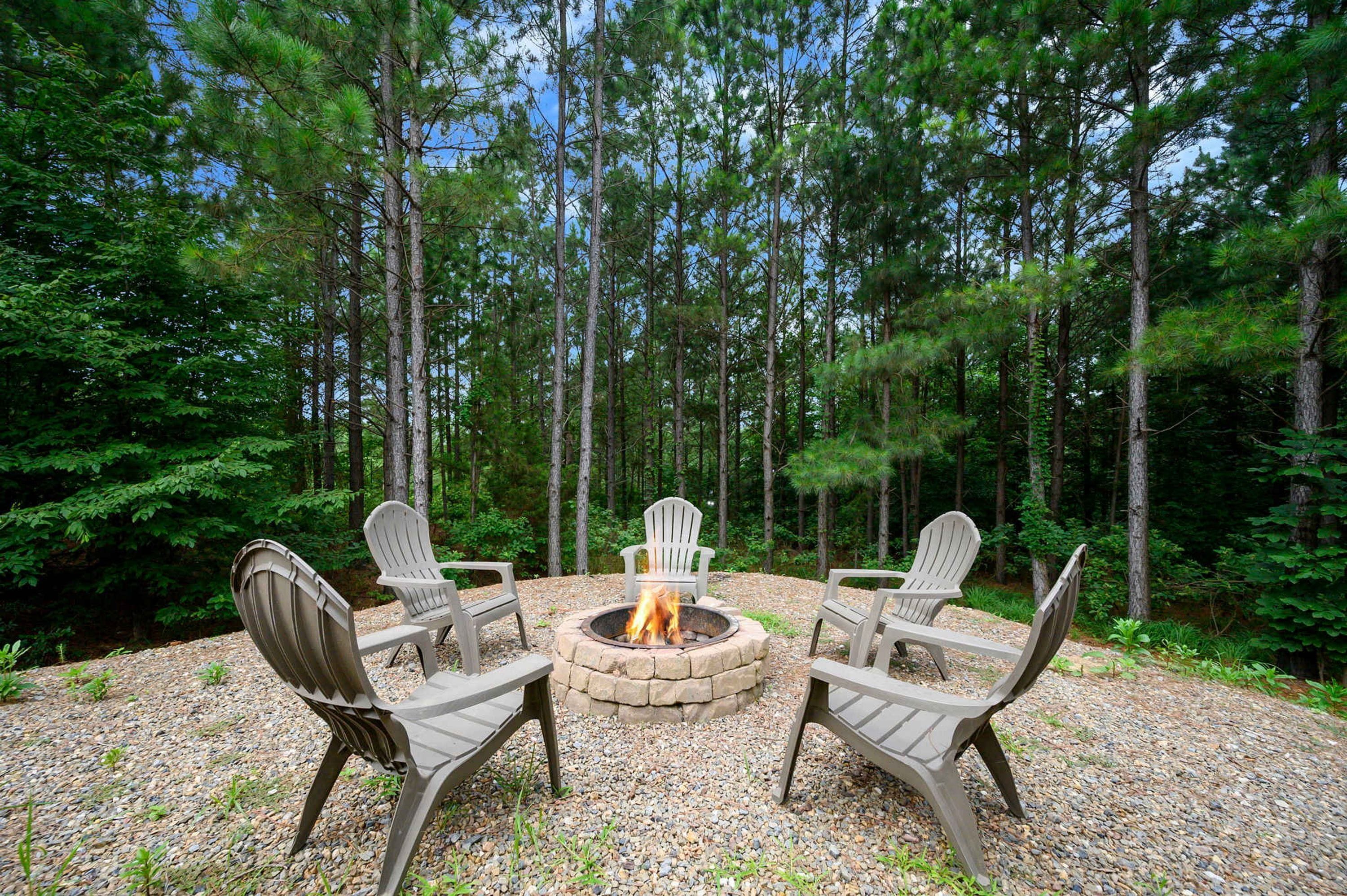 A circular seating arrangement with a central fire pit surrounded by Adirondack chairs in a forest setting.