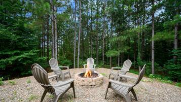 A circular seating arrangement with a central fire pit surrounded by Adirondack chairs in a forest setting.