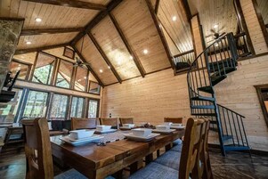 Un comedor amplio con techo alto y vigas de madera, una escalera y ventanas grandes que dejan entrar luz natural.