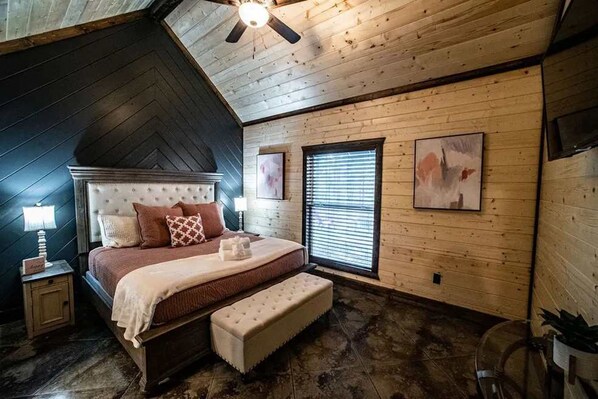 Un dormitorio con techo inclinado, cama con cabecera, una banca, mesita de noche, lámpara y ventana con persianas.