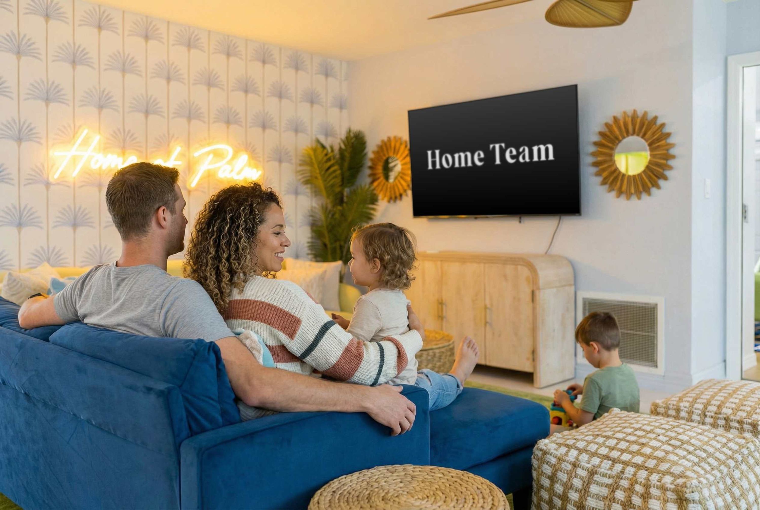 A family on a blue sofa watching TV in a living room with a neon sign reading 'Home Team' and a sunburst decoration.