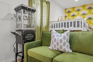 Um sofá verde com uma almofada estampada, uma máquina de pipoca e um berço branco em um quarto com parede florida amarela.